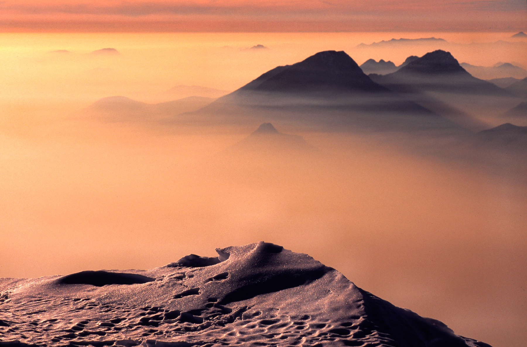 Agosto 2014 - Tramonto invernale dipinto di rosa sulla Pianura Padana, visto dalla cresta meridionale del Monte Baldo. Uno strato di nebbia avvolge la Pianura, e si infila anche nelle vallate del bresciano e sul Lago di Garda. Le vette più alte si ergono verso il cielo come fossero isole in un candido mare.